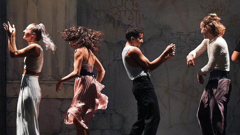 A row of dancing couples on stage in front of two columns and a wall covered in graffiti